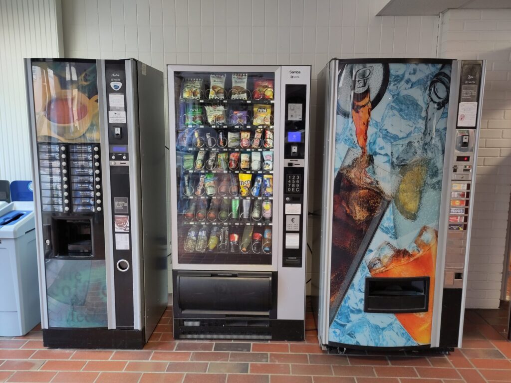 Trois distributeurs automatiques de boissons et de snacks alignés contre un mur blanc dans un espace intérieur.